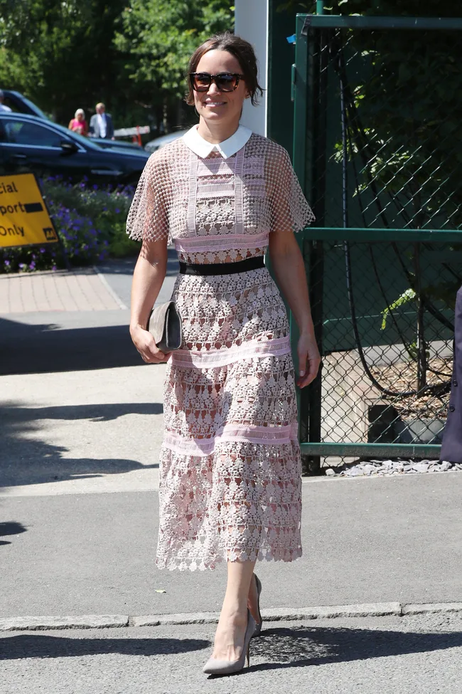 A woman in a lace dress and sunglasses walks outdoors on a sunny day, holding a clutch.