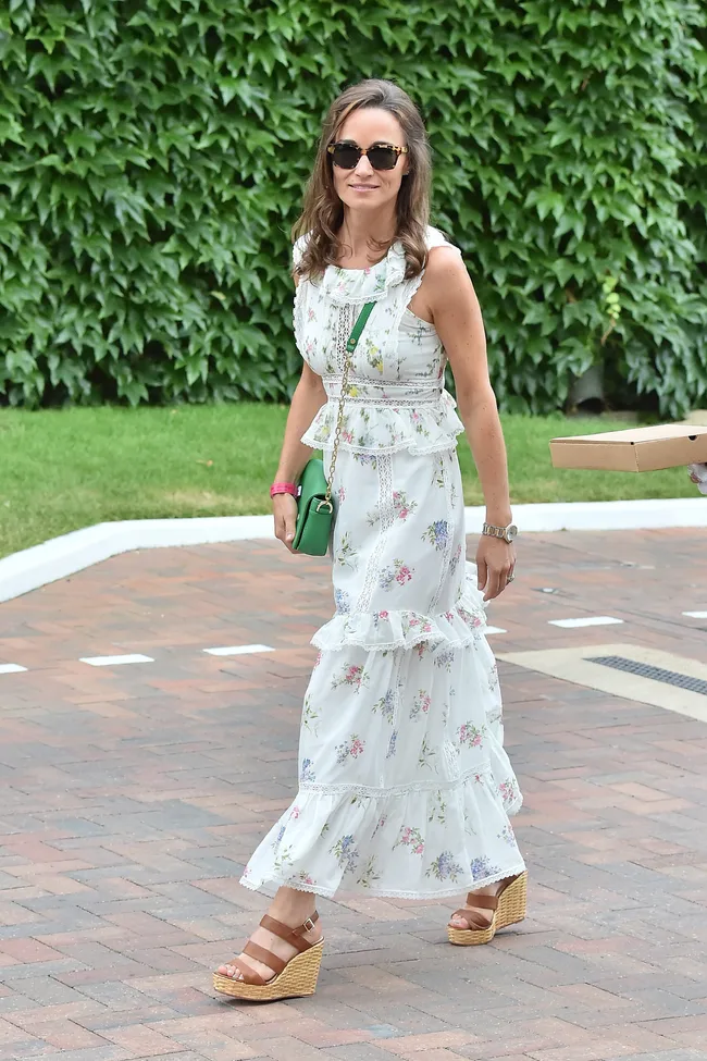 Woman in floral dress and sunglasses walking on brick path with green foliage background.