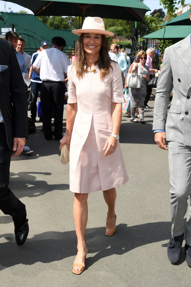 Woman in pink dress and hat walking outdoors, smiling, among a group of people.