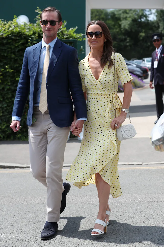 Couple walking outdoors, man in blue blazer and woman in yellow polka dot dress holding hands, both wearing sunglasses.