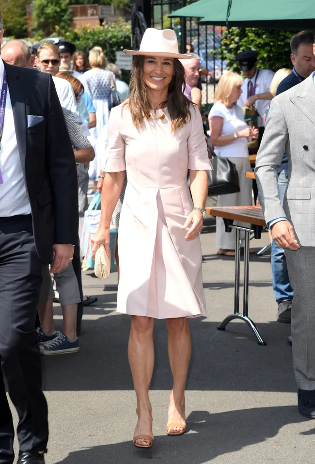 Woman in a pale pink dress and hat walking outdoors, surrounded by people.
