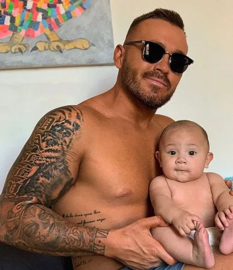 A tattooed man wearing sunglasses holds a baby, sitting indoors with artwork visible in the background.