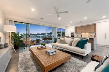 Spacious modern living room with large windows, lake view, white sofas, wooden coffee table, and kitchen in background.