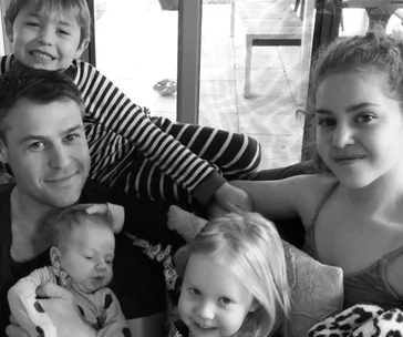 A smiling family with four children, including a baby, sits together on a couch in a cozy setting.