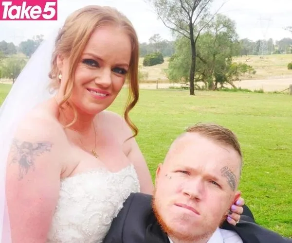 Bride in white dress and groom in suit sitting on grass, smiling outdoors with trees and fields in background.