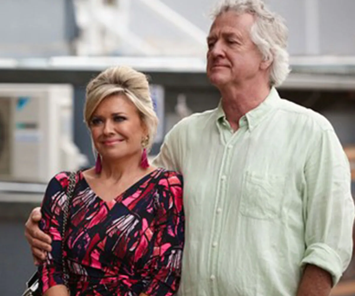 Two actors smiling on set; the woman wears a colorful dress, the man in a light shirt, both appear relaxed and content.