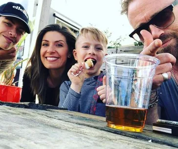 Family smiling with a cup on the table, one child eating, outdoors setting.