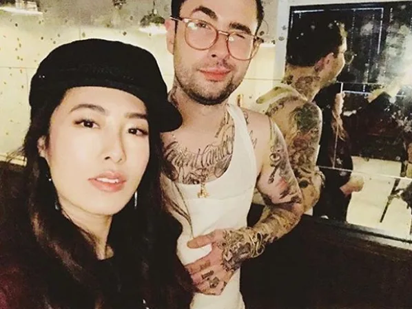 A woman and a tattooed man pose for a casual photo indoors, smiling at the camera.