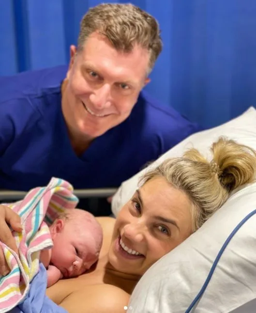 A smiling couple poses with their newborn baby wrapped in a striped blanket in a hospital setting.