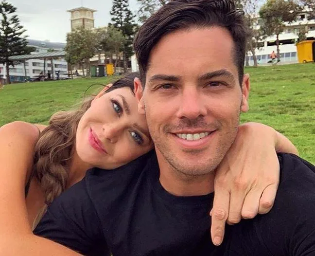 A man and woman smiling, with the woman resting her head on the man's shoulder in a grassy park setting.