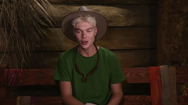 Young person in a green shirt and wide-brimmed hat, sitting indoors on a wooden bench.