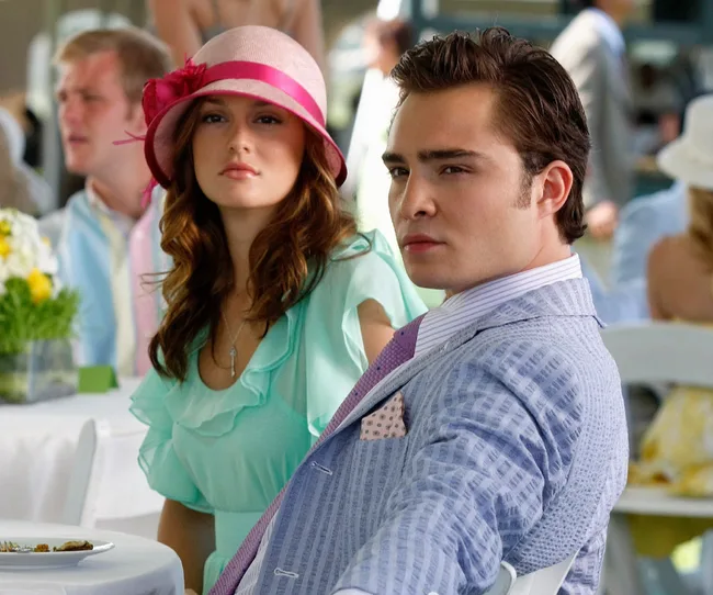 A woman in a pink hat and a man in a suit sitting at an outdoor event, both looking serious.