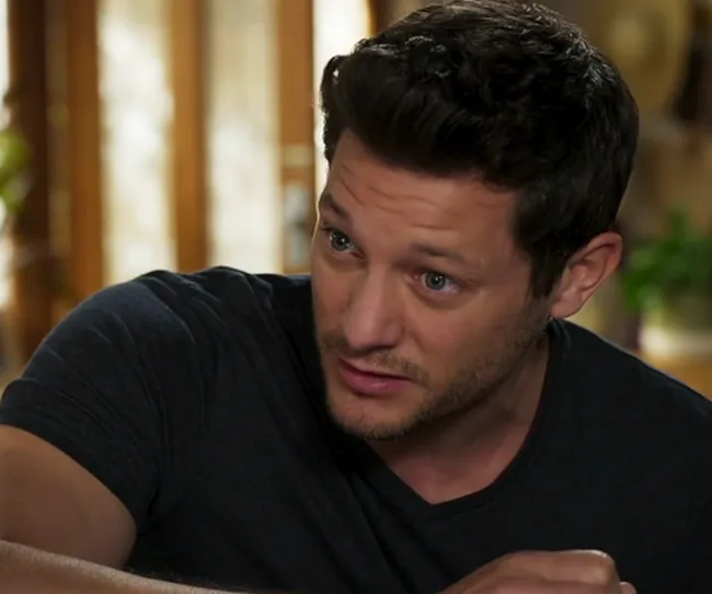 Man with short dark hair, wearing a dark t-shirt, looking slightly to the side indoors with a thoughtful expression.