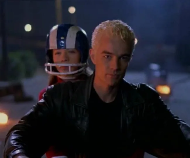Two people riding a motorcycle at night, one wearing a football helmet and the other in a leather jacket.