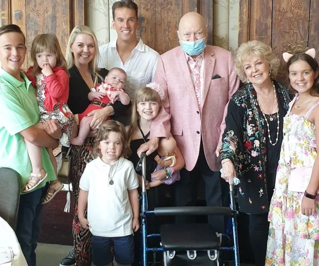 A family portrait with three adults and six children, with one adult using a walker and wearing a pink jacket and mask.