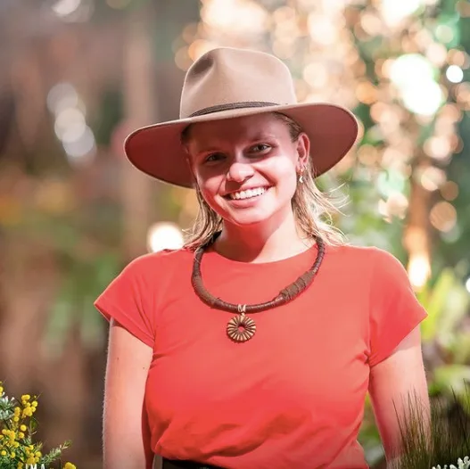 A person in a red shirt and wide-brimmed hat smiles outdoors, surrounded by greenery and bokeh lighting.