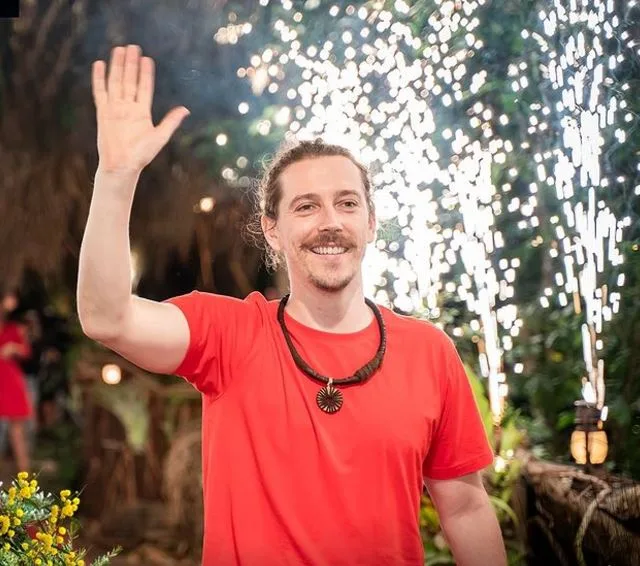 A man in a red shirt waves with fireworks in the background, smiling at an outdoor event.