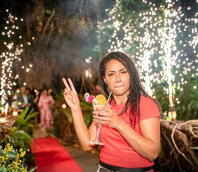 Person in red shirt holding a cocktail and making a peace sign, with festive sparklers and a red carpet in the background.