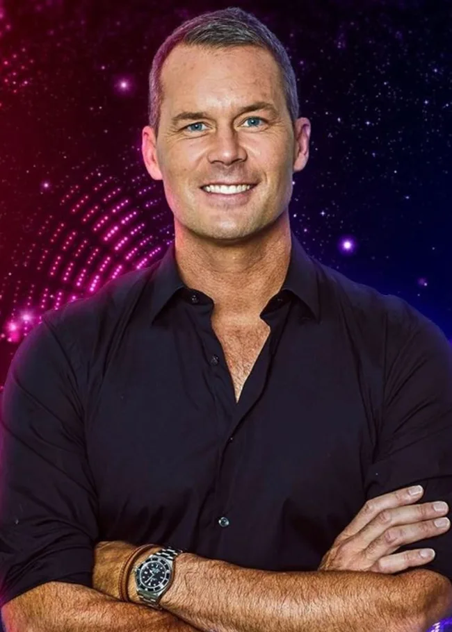 Man in a black shirt smiling with arms crossed, standing against a vibrant pink and blue background.