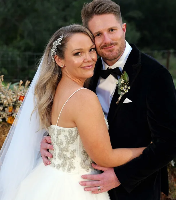 A bride and groom embrace outdoors in formal wedding attire, both smiling at the camera.