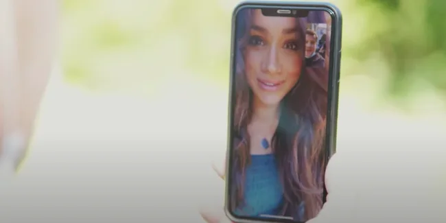 A smartphone showing a video call with a woman in a blue dress, smiling, with a blurred outdoor background.