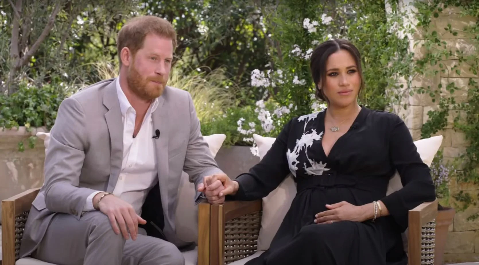 A man and woman sit outdoors, holding hands, surrounded by greenery.