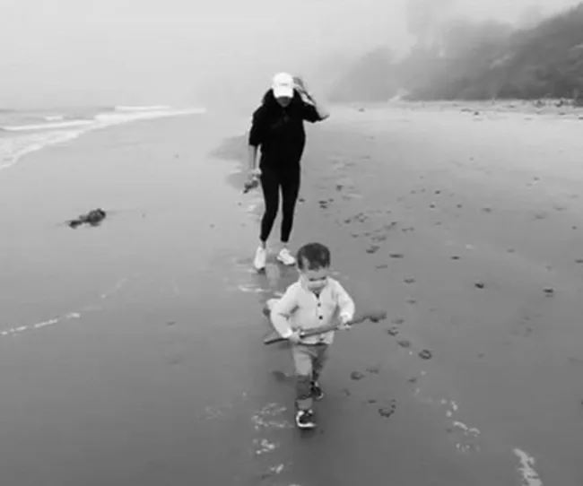 Child holding a stick walks ahead of an adult wearing a hat on a misty beach.