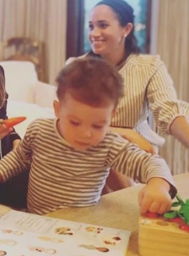 Toddler in striped shirt plays at a table with a woman smiling in the background, indoors.
