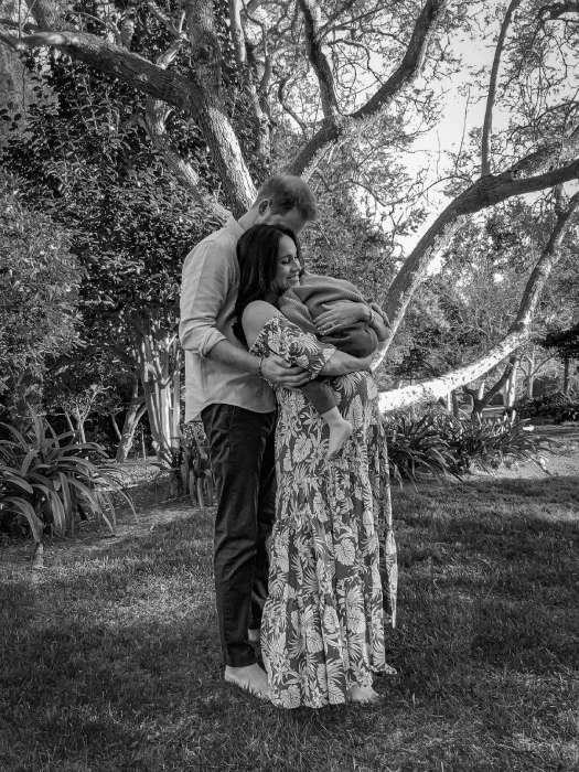 A couple embraces under a tree in a garden, holding a young child wrapped in a blanket.
