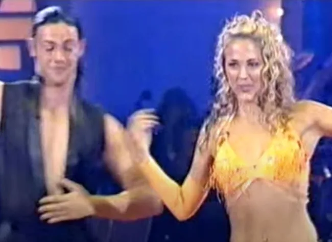 Two people dancing energetically on stage with colorful lights in the background.
