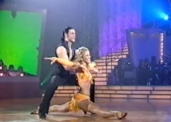 A couple performing a dance routine on a stage with bright lights and a colorful background.