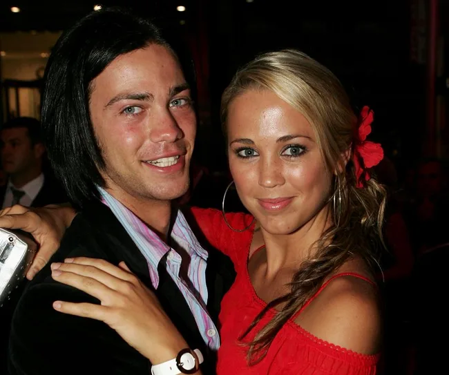 A man and woman smiling at an event, the woman wears a red dress with a matching red flower in her hair.