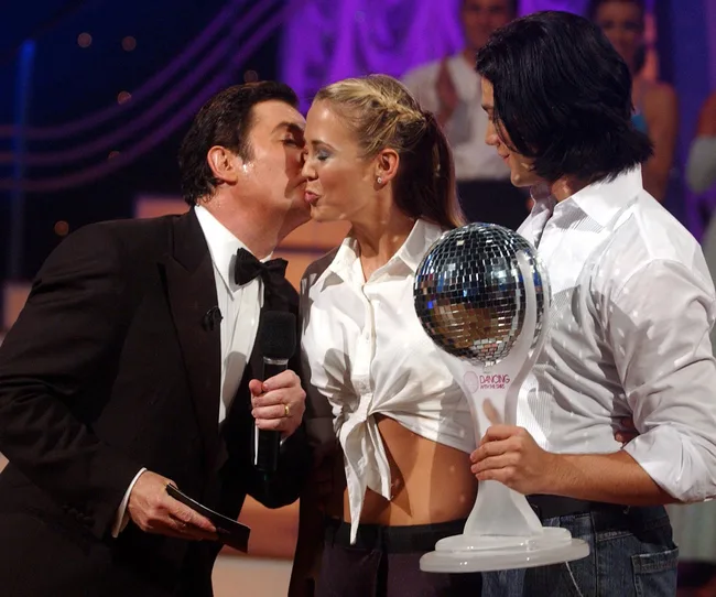 Two people celebrating a victory, holding a "Dancing with the Stars" trophy, while a man in a tuxedo congratulates them.