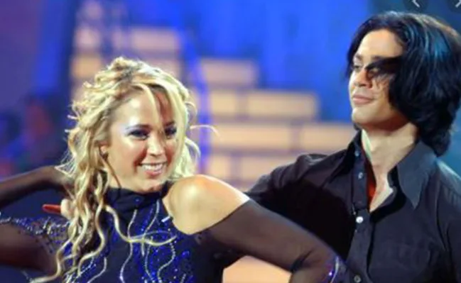 Dancing couple performing on stage, with the woman smiling and the man looking to the side against a blurred background.