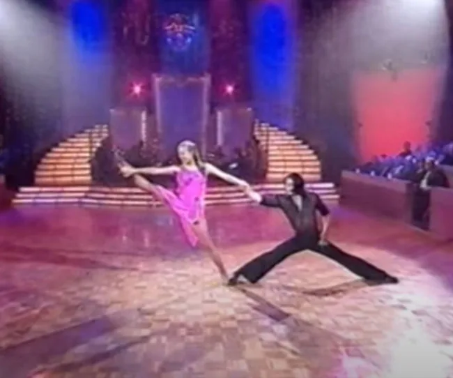 Ballroom dancers performing energetically on a wooden dance floor with colorful stage lights and an audience in the background.