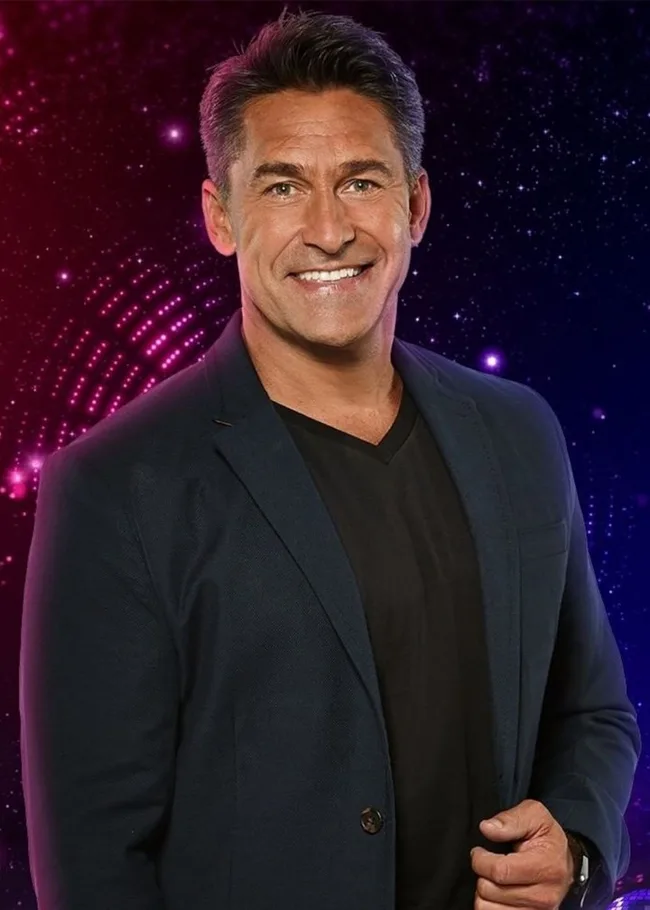 A man in a dark blazer smiles against a colorful, starry background.
