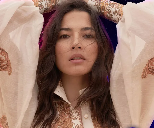 Portrait of a woman with wavy hair, wearing a beige blouse with paisley patterns, hands raised behind her head.