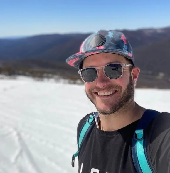 Man smiling in sunglasses and a colorful cap, standing in a snowy landscape with mountains in the background.