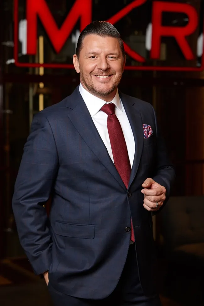 Man in a navy suit with red tie, smiling, standing in front of "MKR" sign.