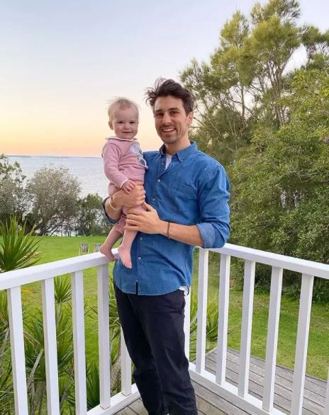 Man holding a baby on a porch, with a scenic view of trees and water in the background during sunset.
