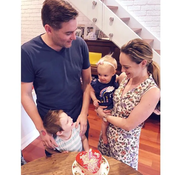 A family with two children stands around a heart-shaped cake with a candle on a wooden table.
