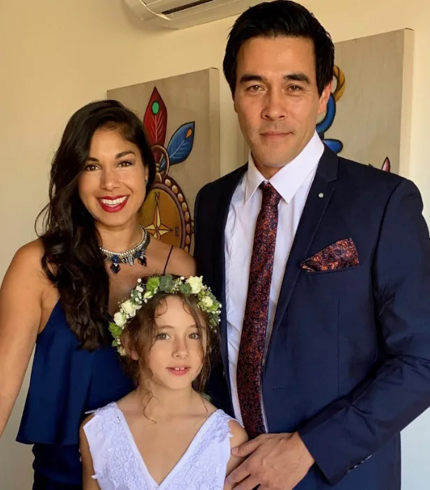 A woman in a blue dress, a man in a suit, and a girl with a flower crown pose together indoors.