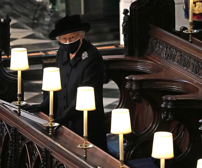 A woman in a black outfit and mask stands in a church pew, surrounded by lamps, during a solemn ceremony.