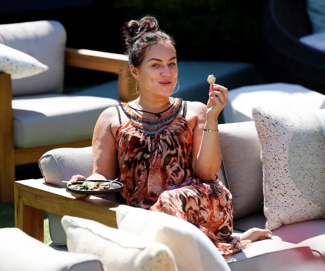 A woman in a patterned dress sits on an outdoor sofa, holding food and smiling, with cushions and chairs around.
