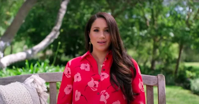 Woman in a red floral dress sitting outdoors on a wooden bench, surrounded by greenery.