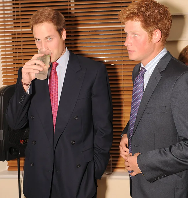 Two men in suits, one drinking from a glass, indoors with blinds in the background.