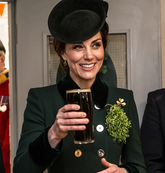 Woman in a green outfit and hat holding a pint of dark beer, smiling with shamrock accessory.