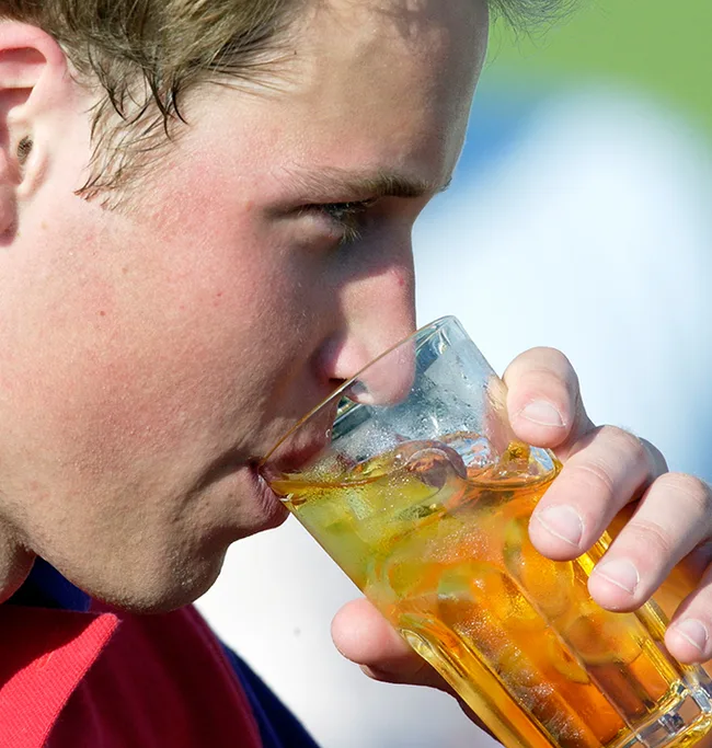 Young man drinking from a glass of iced tea or similar beverage, outdoors in a sunny setting.