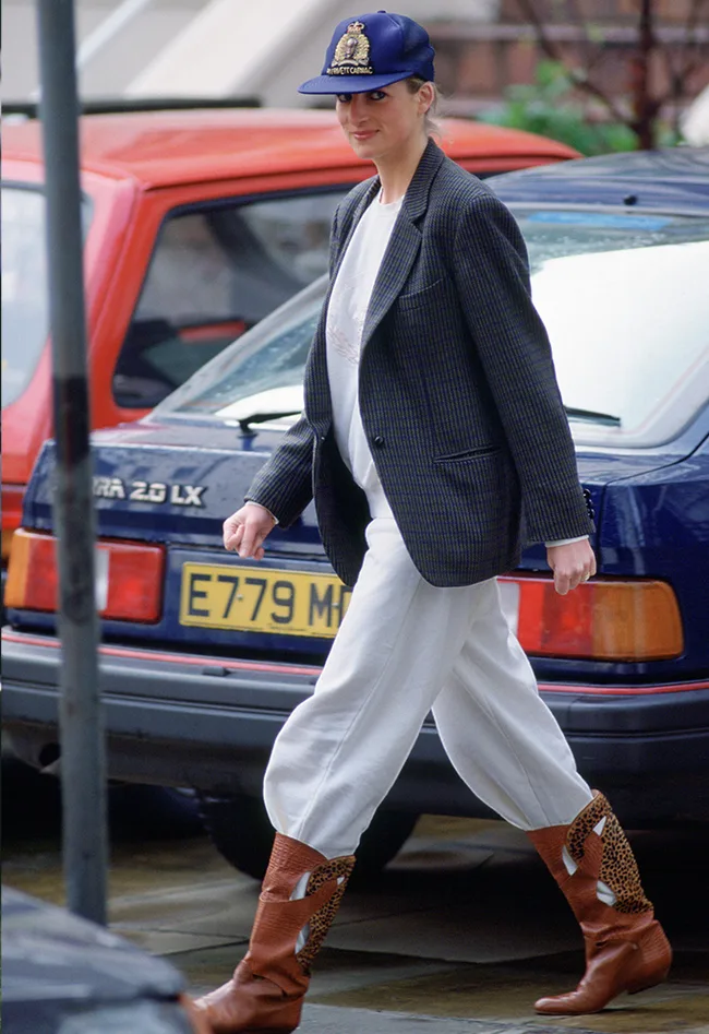 A person in a blazer, white pants, brown boots, and a blue cap walking past parked cars.