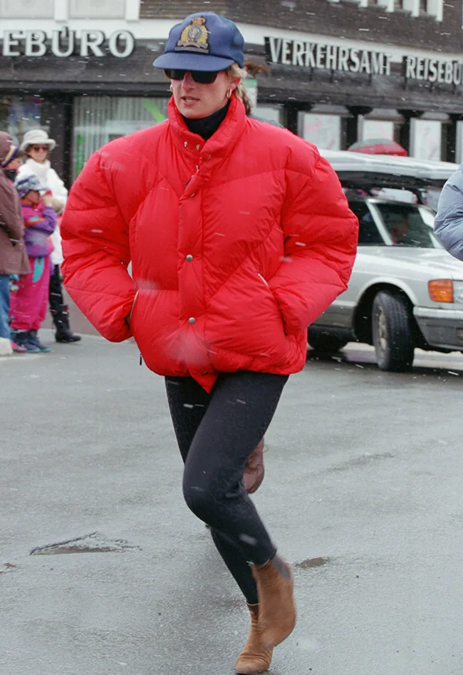 A person in a red puffer jacket, black leggings, brown boots, sunglasses, and a blue cap walking on a snowy street.